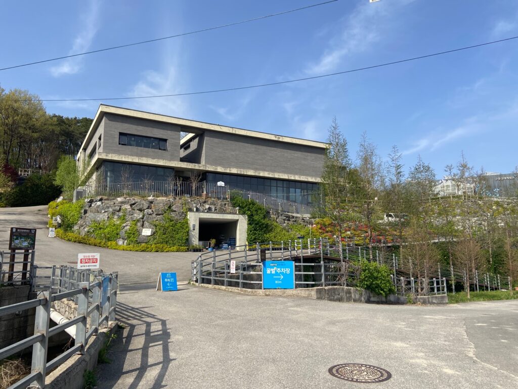 Modern gray two-story building on a rocky hillside, with a driveway and ramp leading to an underground entrance beneath a landscaped terrace.