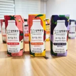 나무 테이블 위에 나란히 놓인 상하목장 유기농 주스 3종 팩 — 왼쪽부터 사과딸기케일, 사과오렌지케일, 사과블루베리케일, 각 125mL 설탕무첨가 표기