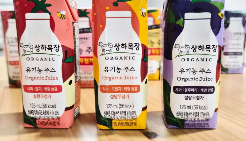 나무 테이블 위에 나란히 놓인 상하목장 유기농 주스 3종 팩 — 왼쪽부터 사과딸기케일, 사과오렌지케일, 사과블루베리케일, 각 125mL 설탕무첨가 표기